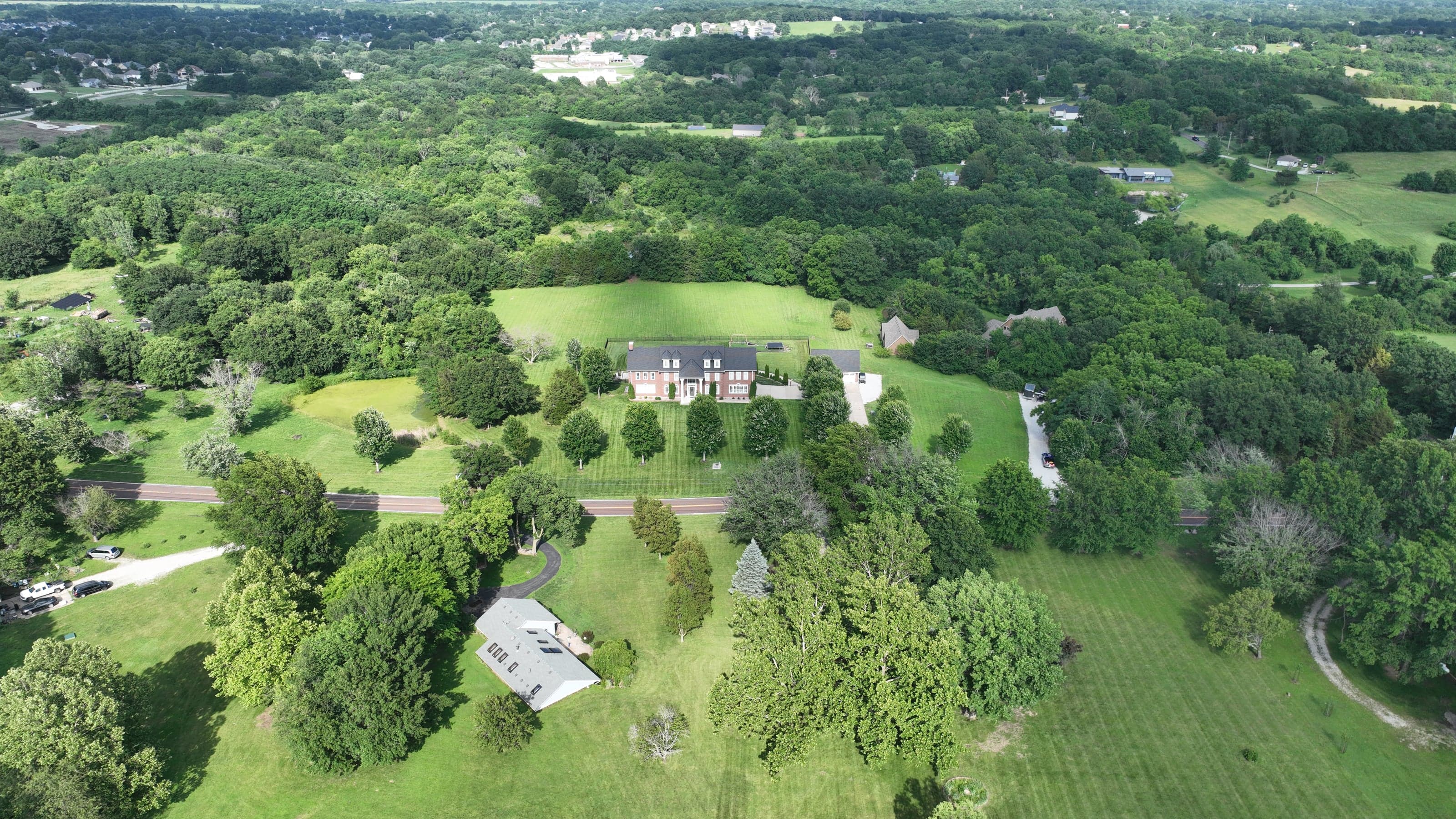 Aerial drone photo of a brick estate home with a pool and circular driveway on a wooded lot